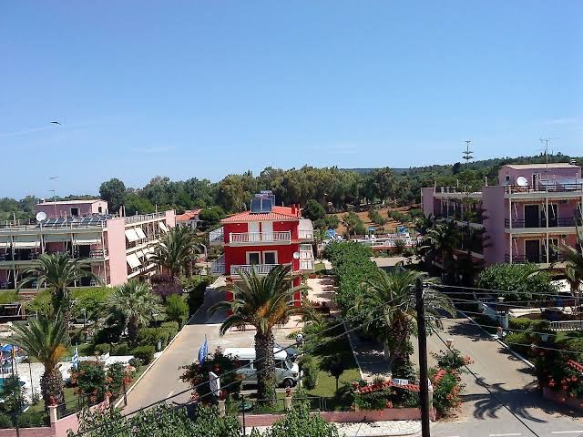 Panorāmas skats uz Brati Arkoudi Hotel kompleksu Peloponēsā — rozā un sarkanās ēkas, palmas un apkārtnes apstādījumi