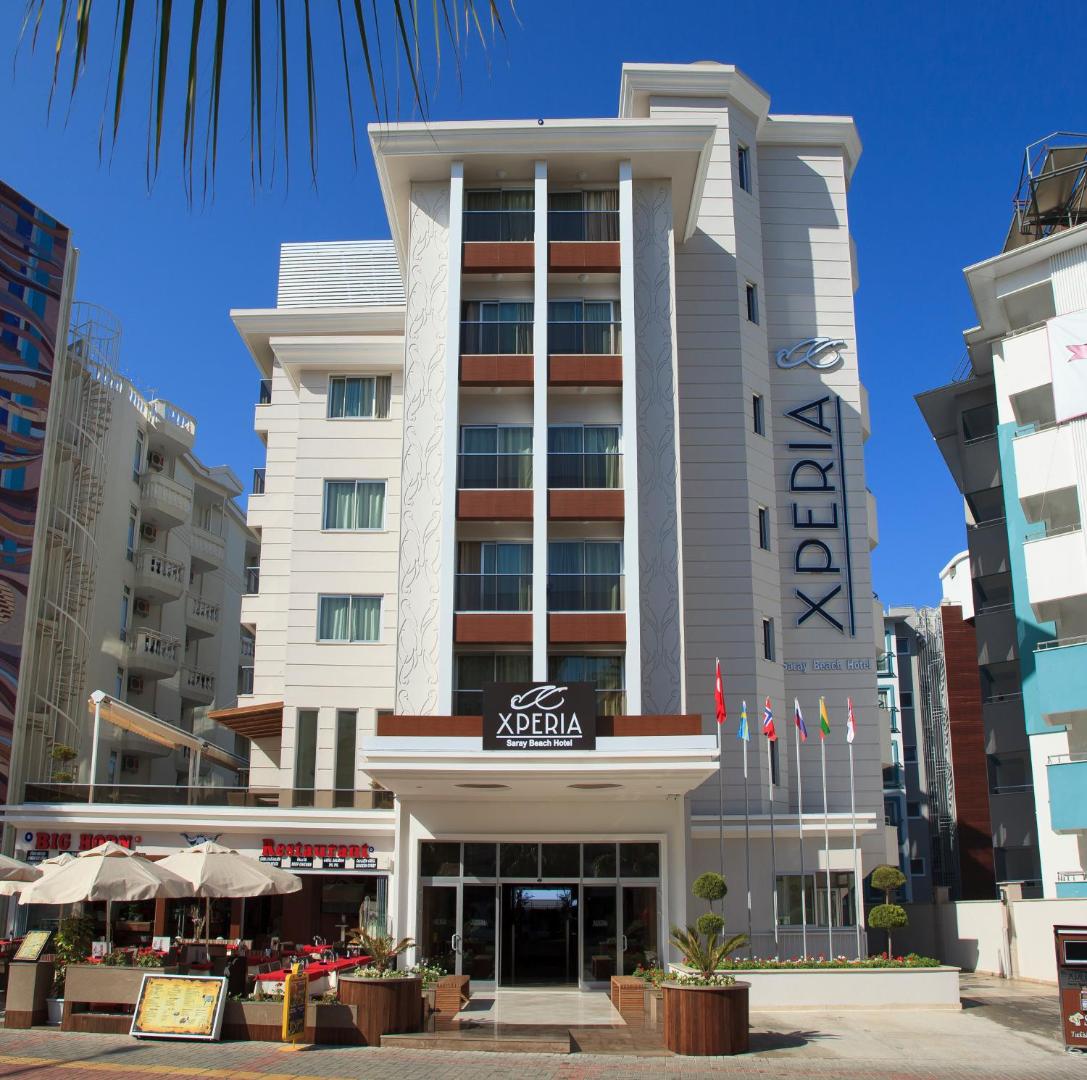 Xperia Saray Beach Hotel galvenā ēka Alānijā dienas laikā — balta daudzstāvu viesnīca ar balkoniem un karogiem pie ieejas