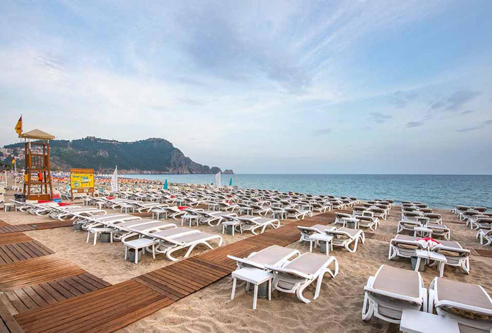 Xperia Saray Beach viesnīcas privātā pludmale Alanjā ar daudziem sauļošanās krēsliem, koka laipām un skatu uz Alanjas cietokšņa kalnu