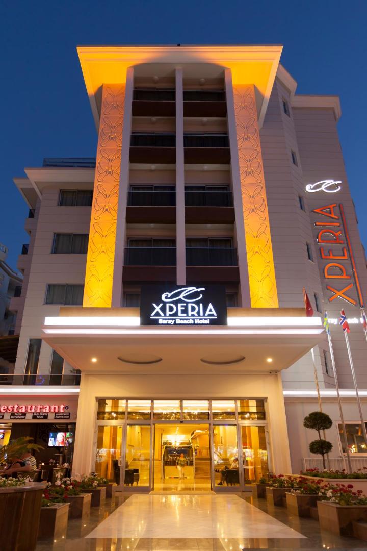 Xperia Saray Beach Hotel ieeja Alānijā vakara apgaismojumā — moderna fasāde ar izgaismotu logo un stikla durvīm piekrastes kūrortā