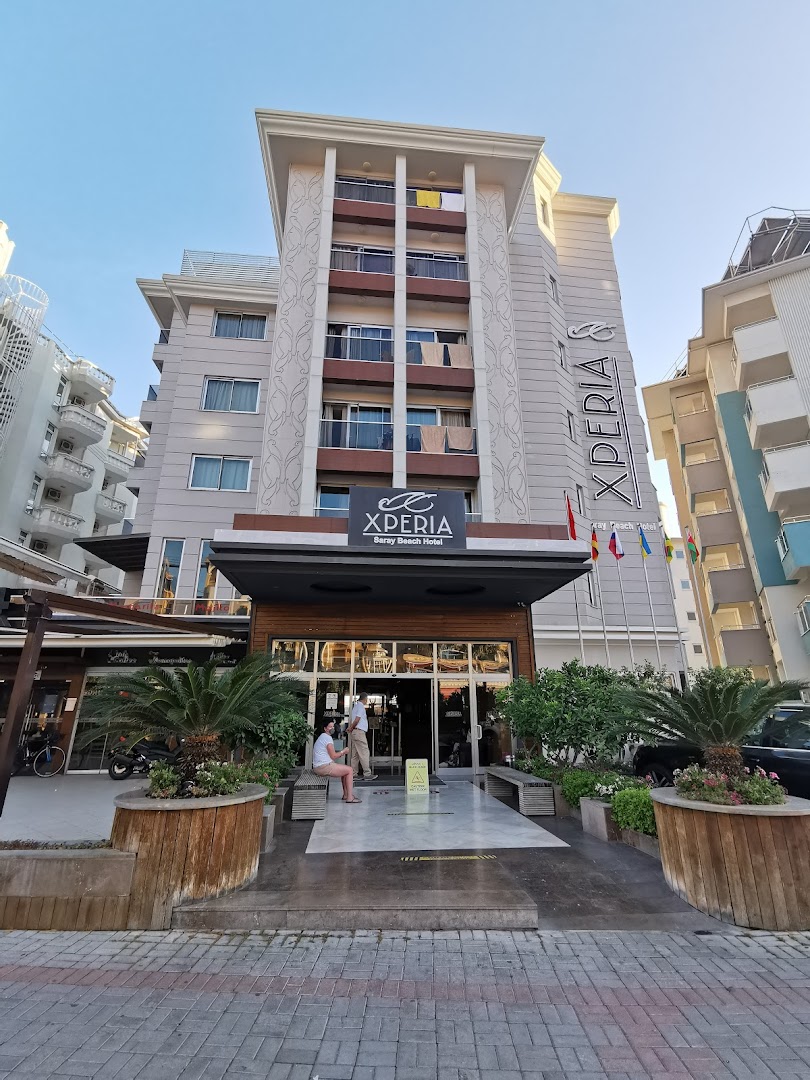 Xperia Saray Beach Hotel fasāde no ielas puses — ieeja ar palmām koka podos un modernu arhitektūru Alānijas pilsētvidē