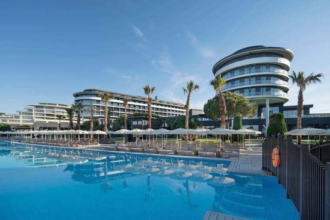 Voyage Belek Golf & Spa galvenais āra baseins ar saulessargiem un modernu viesnīcas arhitektūru Belekas reģionā, Turcijā