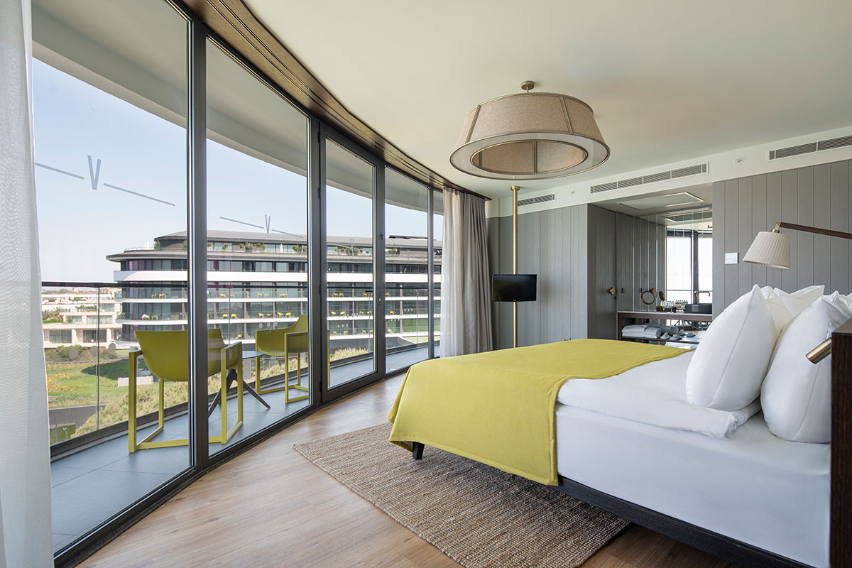 Voyage Belek Golf & Spa moderns numurs ar panorāmas logiem, skatu uz viesnīcas korpusu un mēbelētu balkonu Antaljas reģionā