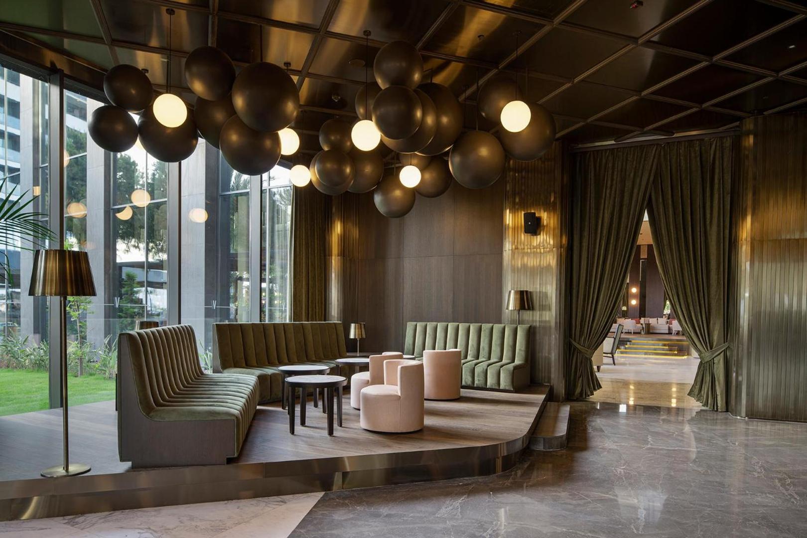 Voyage Belek Golf & Spa lobby bāra atpūtas zona ar zaļiem dīvāniem, dizaina lampām un skatu uz dārzu