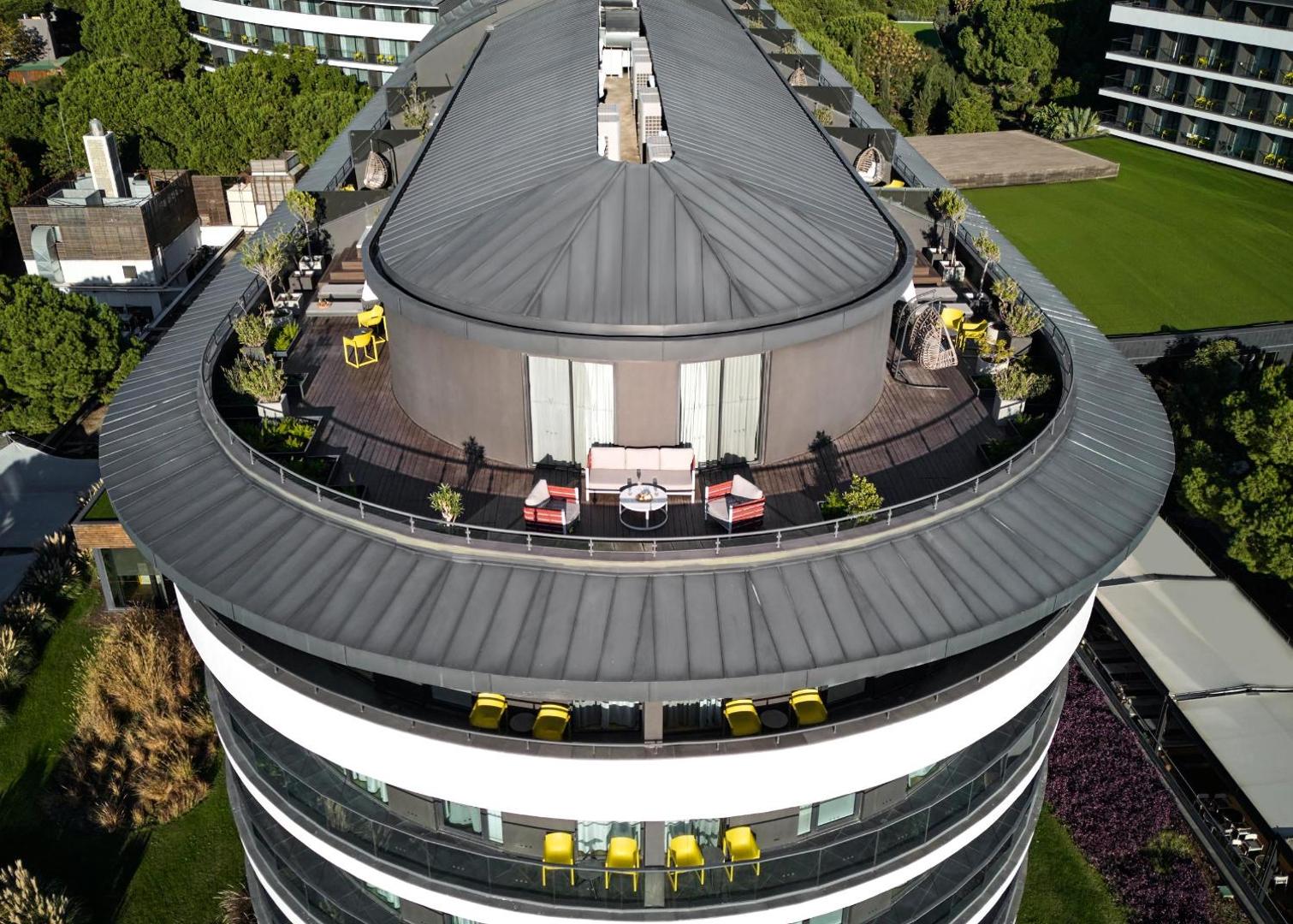 Voyage Belek Golf & Spa jumta terase ar atpūtas mēbelēm un skatu uz priežu mežu Belekā, Turcijā — moderna arhitektūra no putna lidojuma