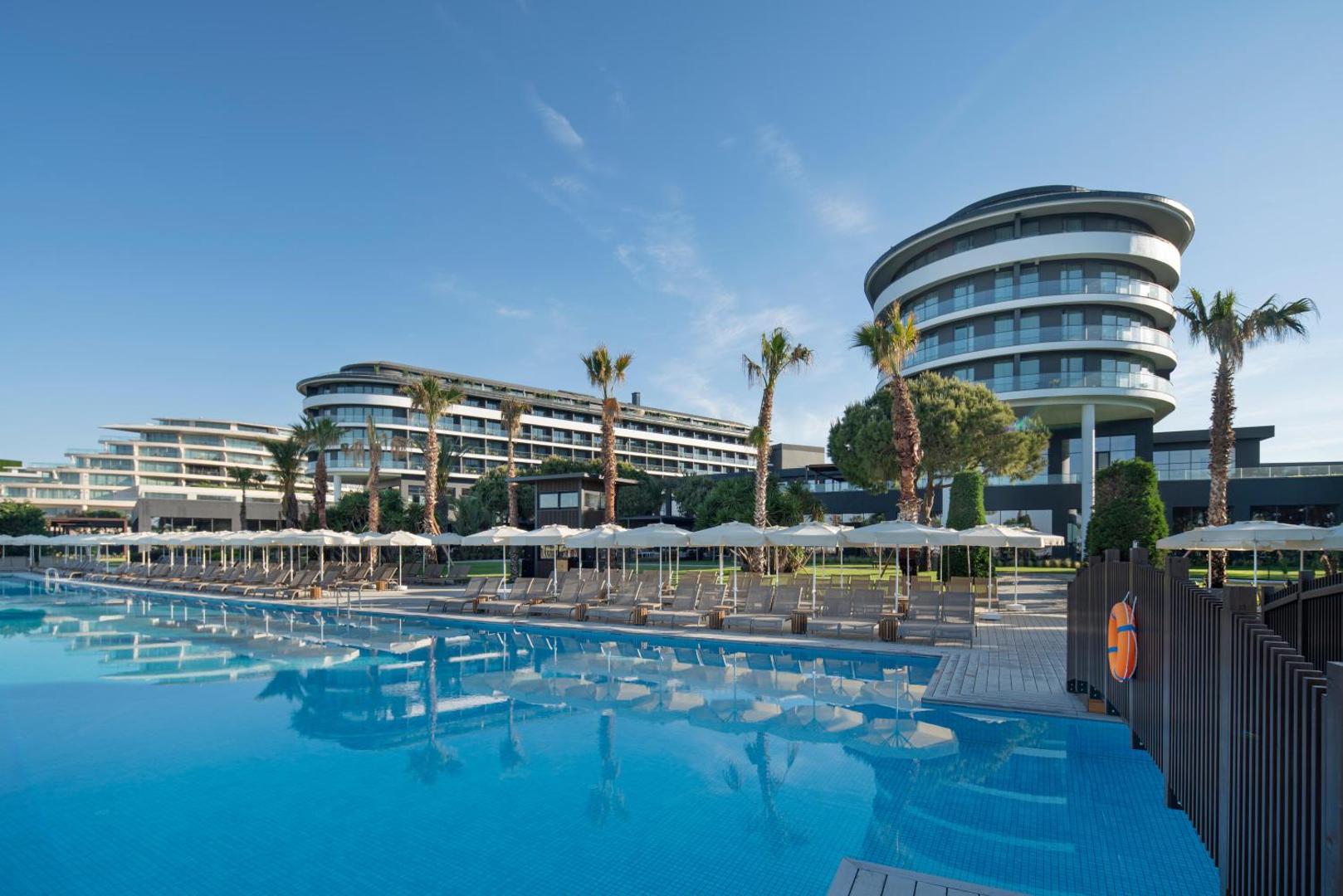 Voyage Belek Golf & Spa galvenais āra baseins ar atpūtas krēsliem, saulessargiem un viesnīcas ēkām Belekā, Turcijā