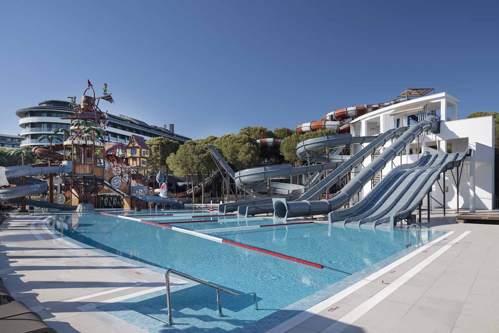 Voyage Belek Golf & Spa plašais āra baseins ar vairākiem lielajiem ūdens slidkalniņiem un viesnīcas korpusu fonā Belekā