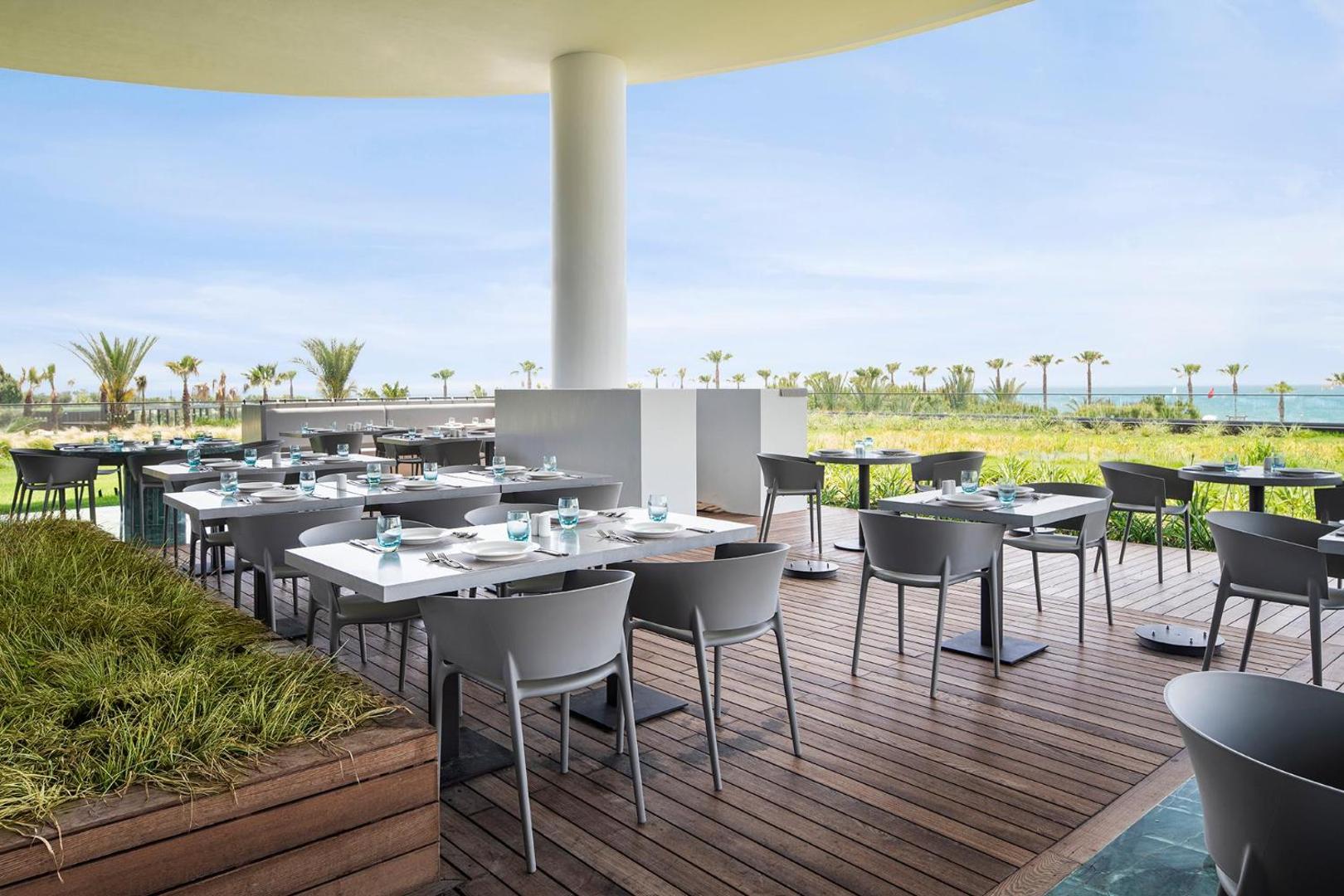 Voyage Belek Golf & Spa āra terase ar skatu uz jūru un palmām, klāti galdi un koka grīdas segums