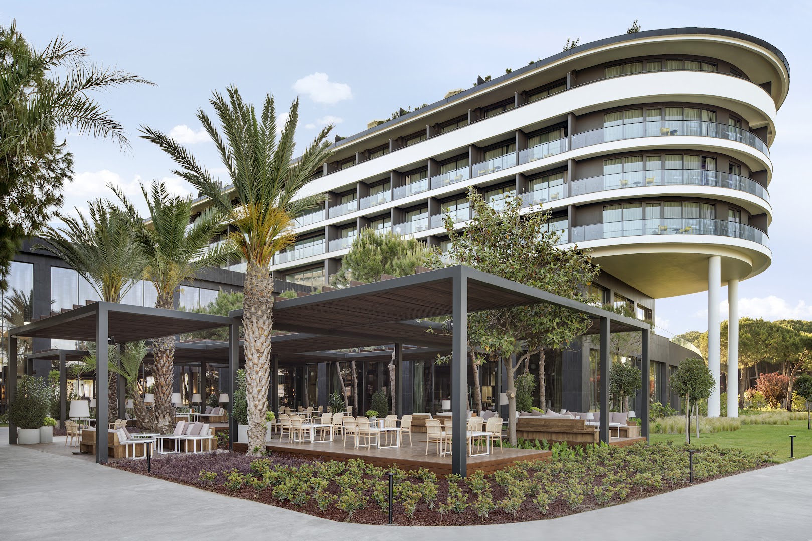 Voyage Belek Golf & Spa viesnīcas fasāde un āra restorāna terase ar palmām un dārzu Belekas kūrortā — saulains dienas skats