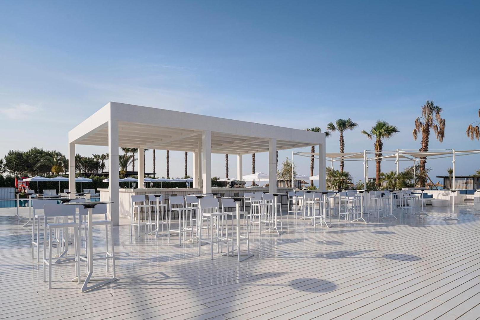 Voyage Belek Golf & Spa āra terases bārs ar baltām mēbelēm, koka klāju un palmām fonā saulainā dienā Belekā