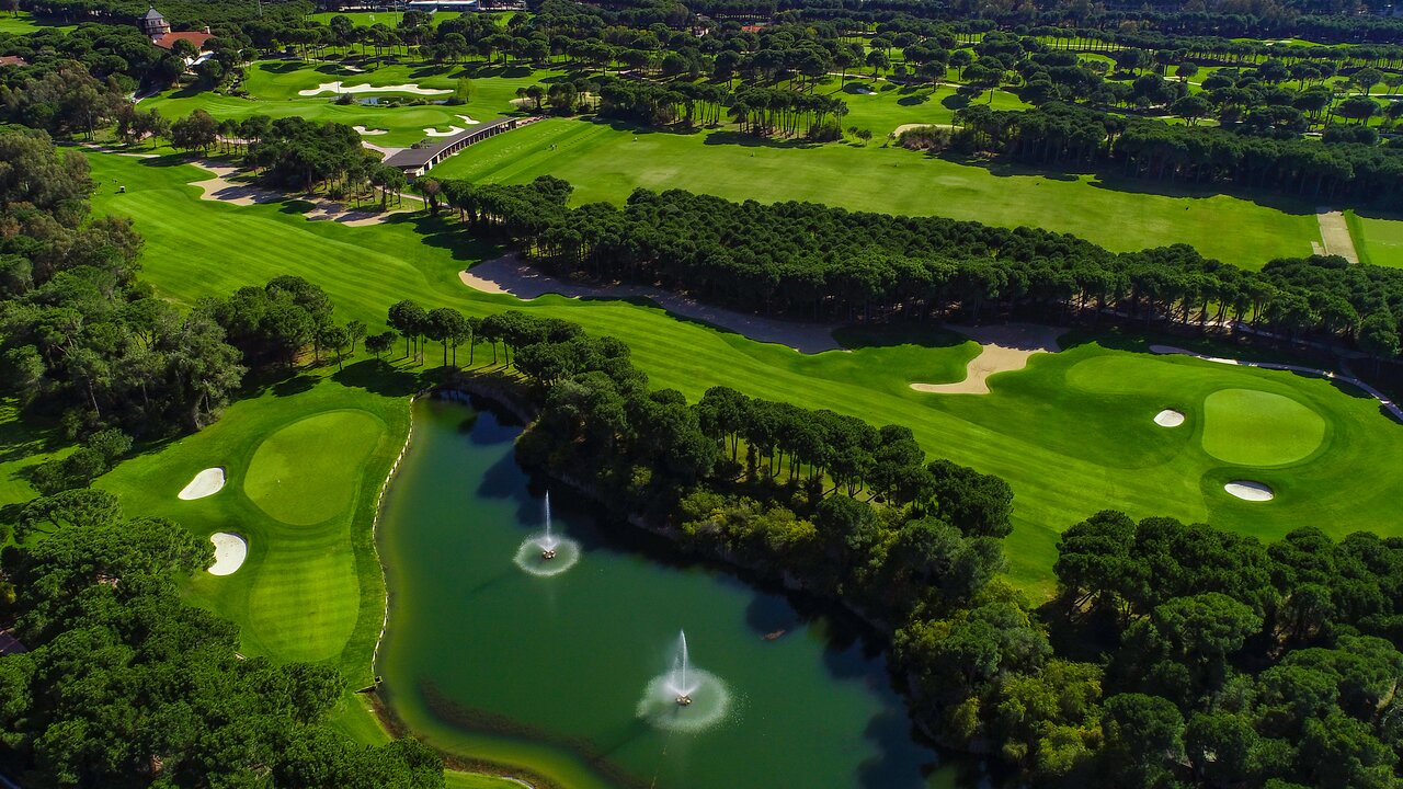 Aerofoto skats uz Voyage Belek Golf & Spa plašajiem golfa laukumiem, priežu mežu un ezeru ar strūklakām Belekā