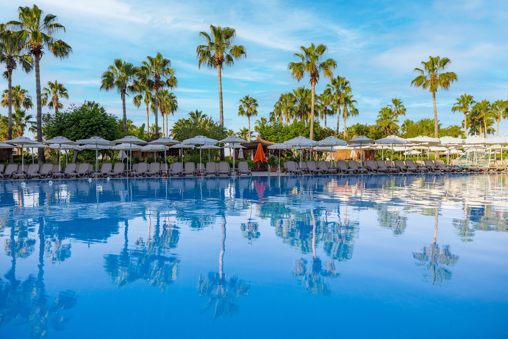 VONRESORT Golden Coast baseins ar palmu atspulgiem ūdenī, rindu ar saulessargiem un zilu debesu fonu Turcijas piekrastē