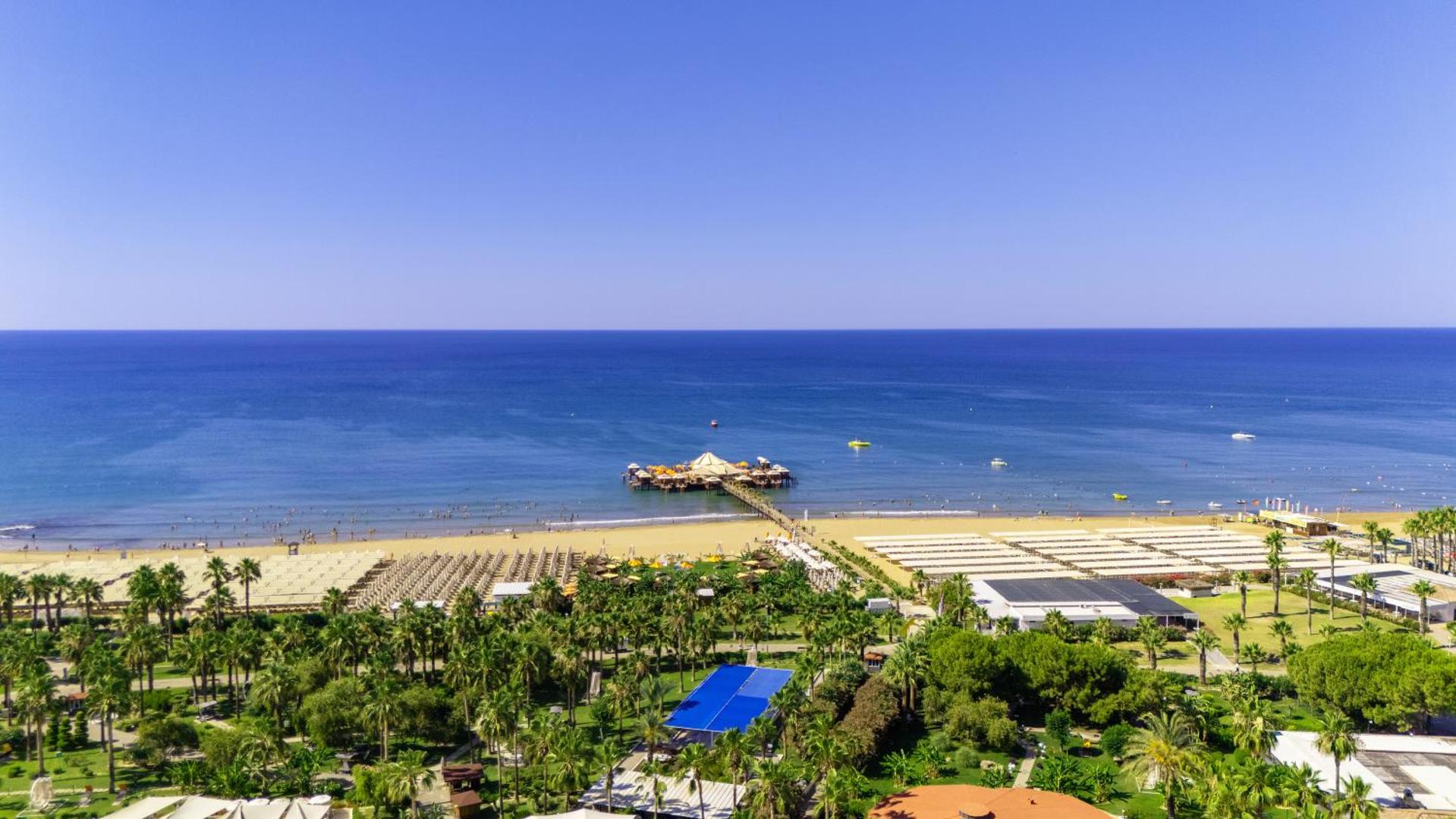 Von Resort Golden Coast panorāmas skats uz plašo smilšu pludmali, zilo Vidusjūru un koka piestātni ar atpūtas zonu Sidē, Turcijā.