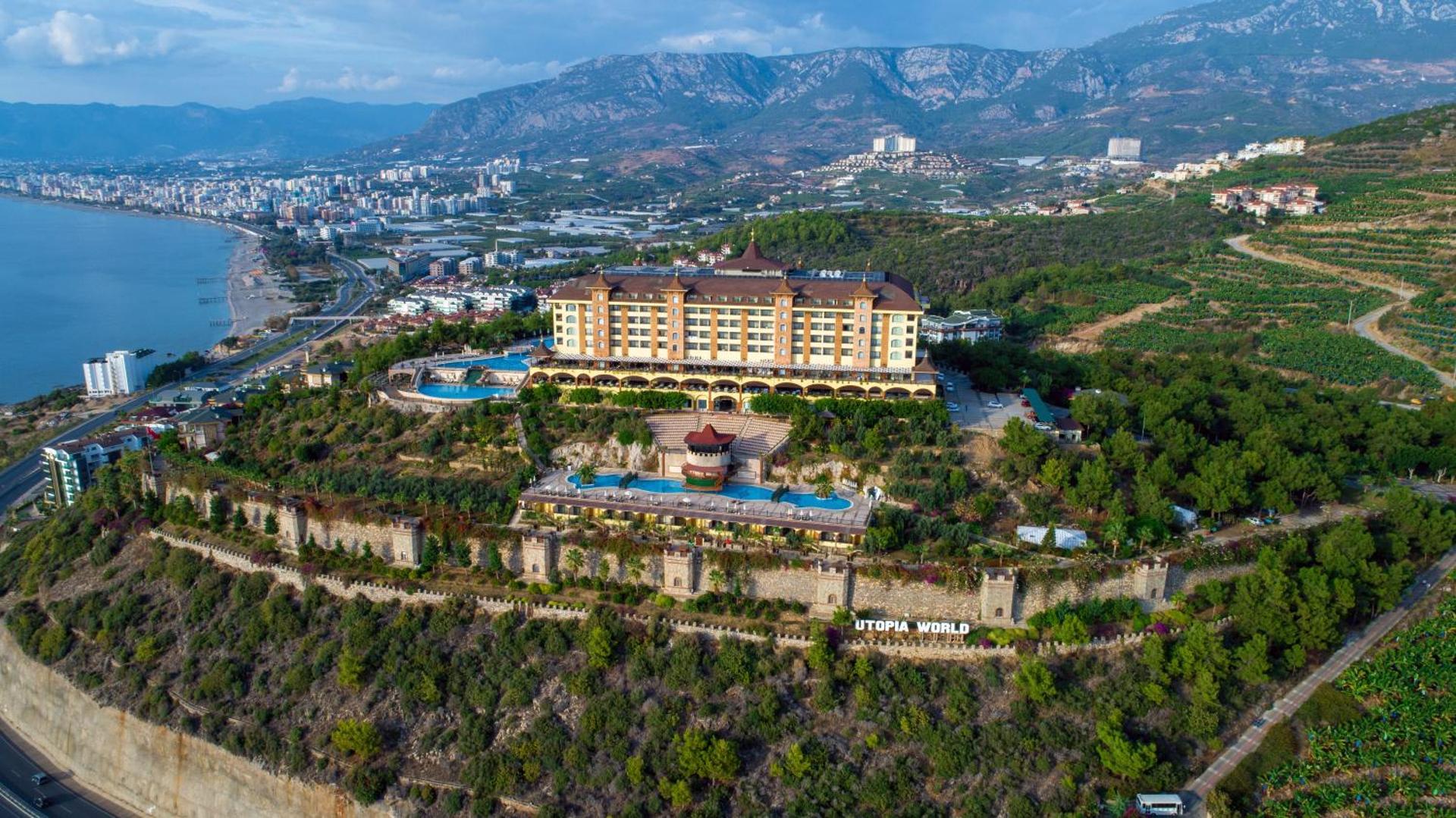 Utopia World Hotel galvenā ēka un āra baseini uz kalna fona Alanjā — arhitektūra, sauļošanās terases un zaļā zona
