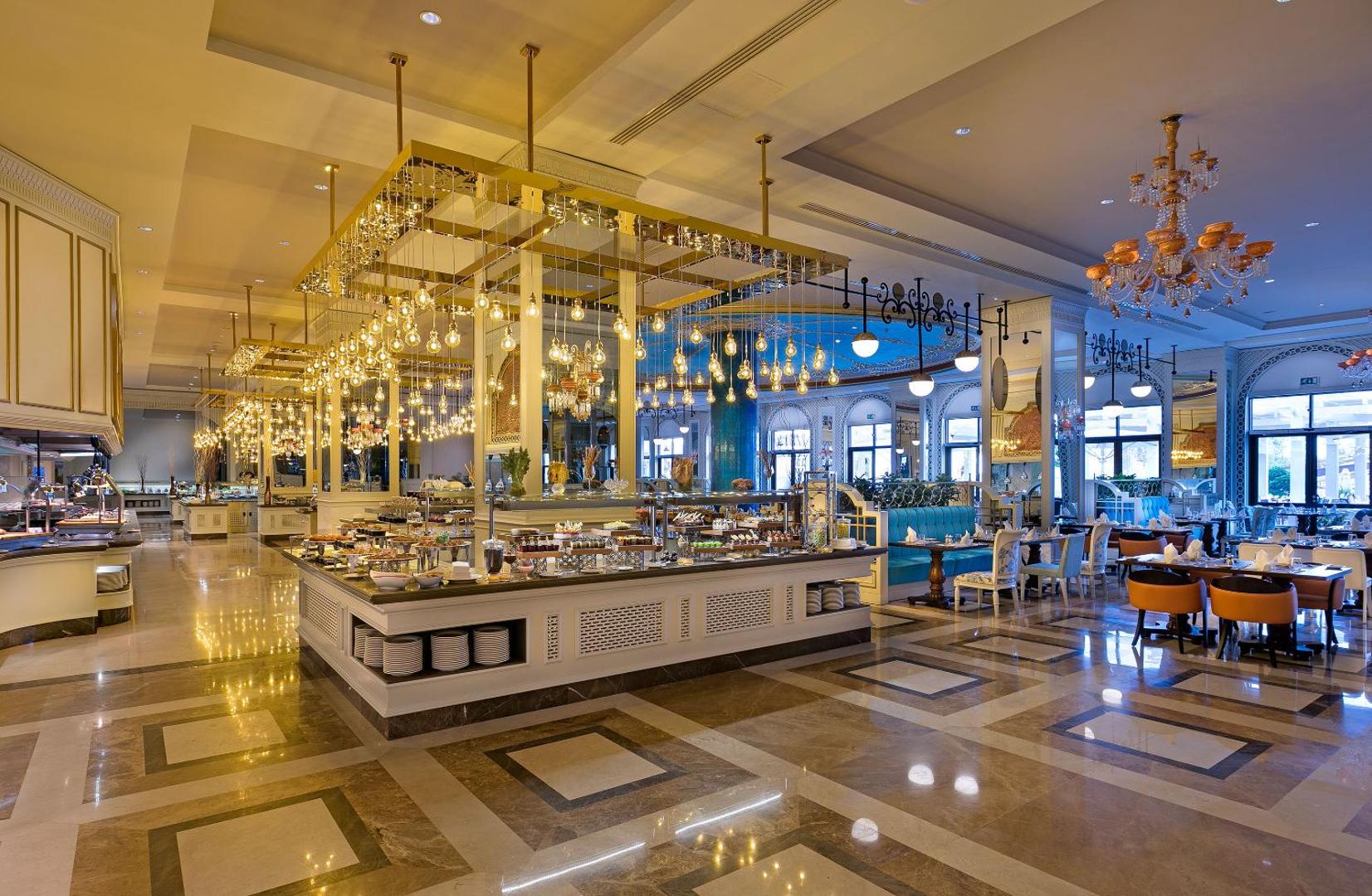 Gaišs Titanic Mardan Palace bufetes restorāns ar greznām lustrām, ziliem dīvāniem un plašu desertu izvēli uz centrālās letes