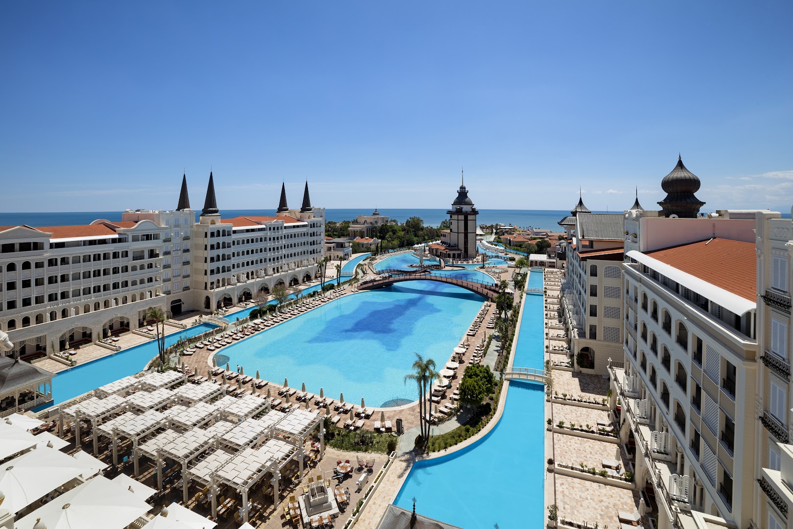 Titanic Mardan Palace centrālais baseins ar tiltiņiem un sauļošanās krēsliem saulainā dienā Antālijas reģionā