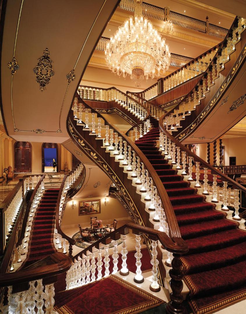 Titanic Mardan Palace monumentālās iekštelpu kāpnes ar sarkanu paklāju, stikla margām un iespaidīgu lustru augšpusē, demonstrējot pils arhitektūru