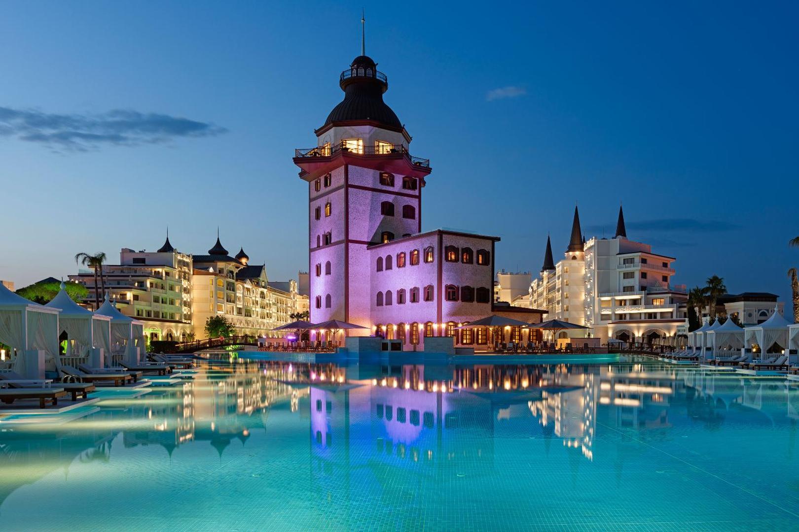 Titanic Mardan Palace viesnīcas centrālais baseins un vēsturiskā stila tornis vakarā Antaljas reģionā, Turcijā — izgaismota arhitektūra un atpūtas zonas.