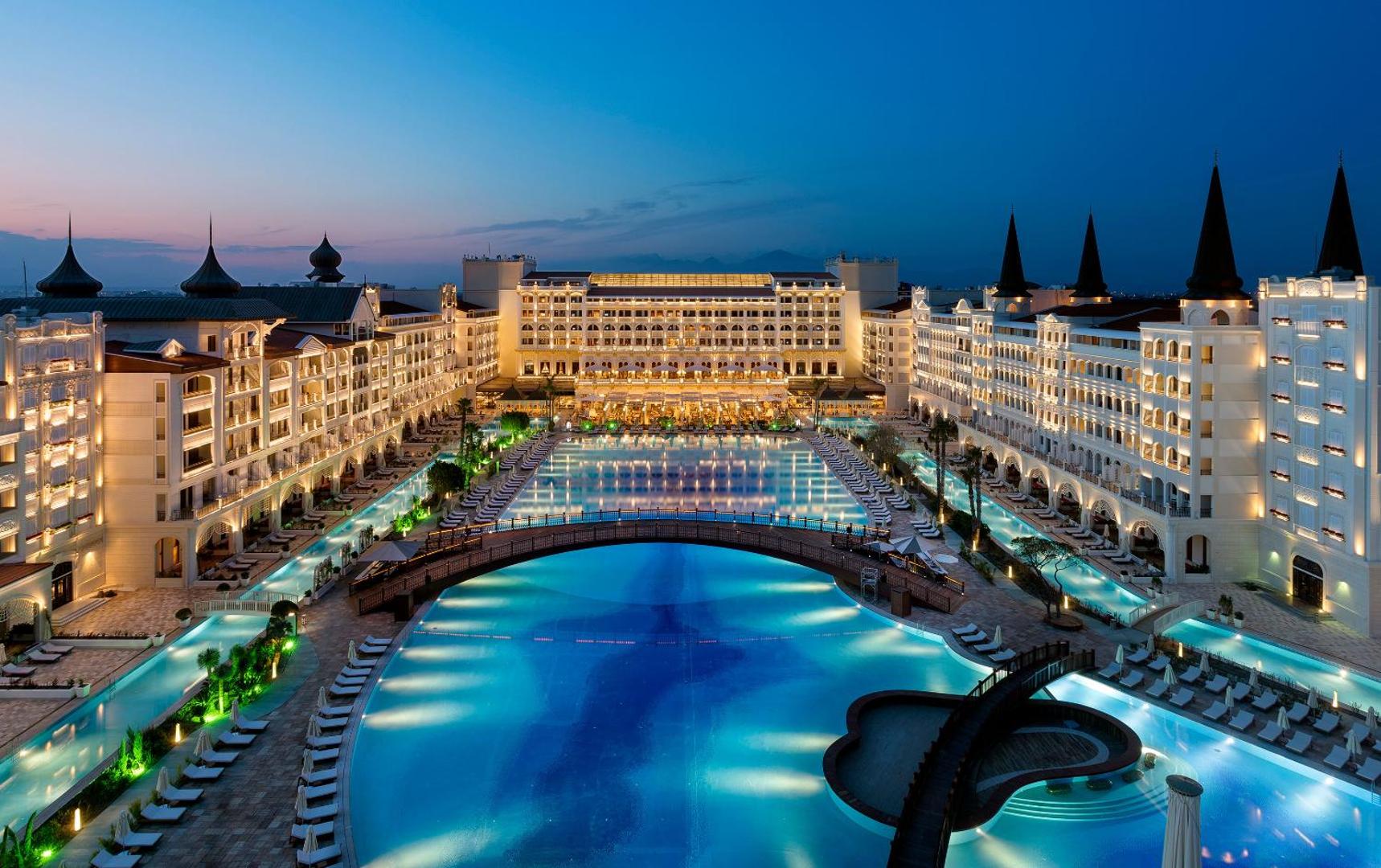 Titanic Mardan Palace panorāmas skats vakarā uz plašo baseinu kompleksu, tiltu un viesnīcas korpusiem ar greznu apgaismojumu Antaljā.