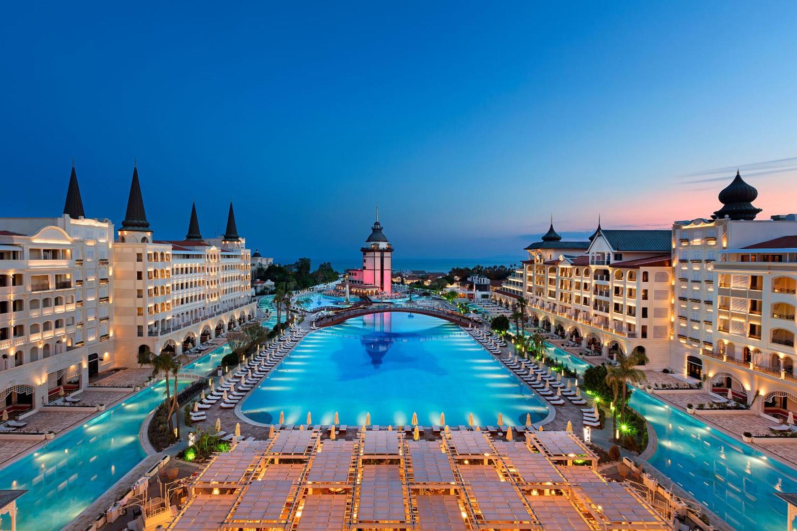 Skats no augšas uz Titanic Mardan Palace centrālo baseinu, tiltu un viesnīcas ēkām saulrieta laikā — plaša teritorija un atpūtas terases.