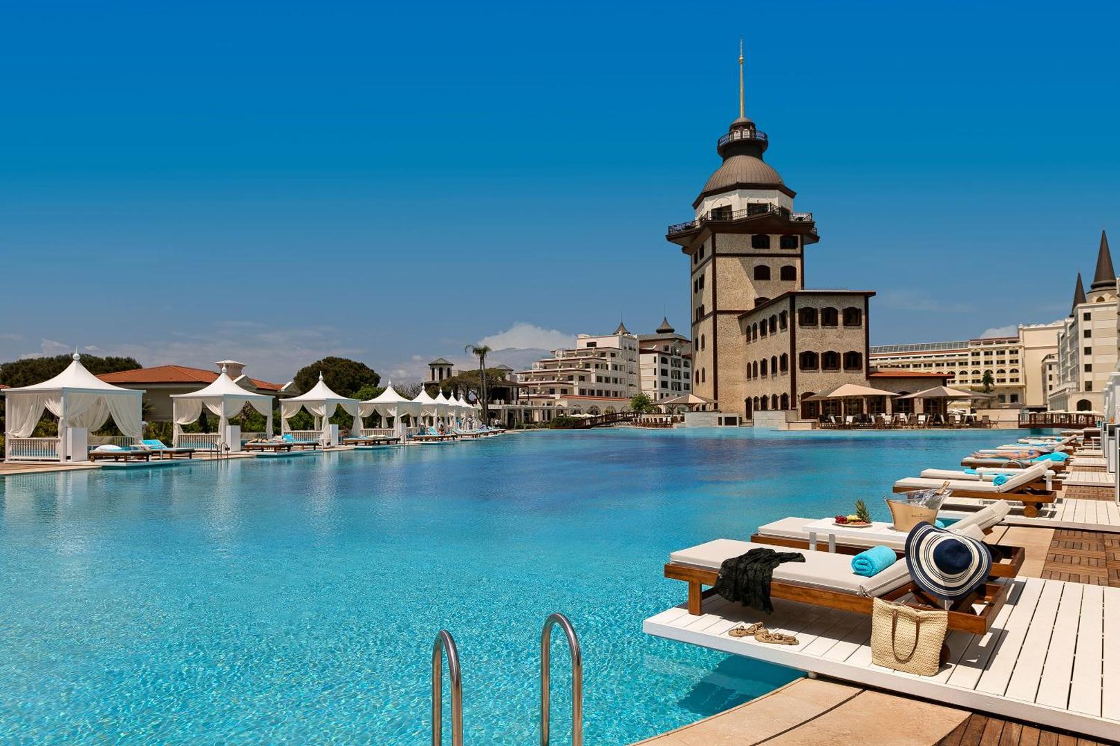 Titanic Mardan Palace āra baseins ar sauļošanās krēsliem un baltām teltīm dienas laikā — skats uz viesnīcas raksturīgo torni un zilo debesi.