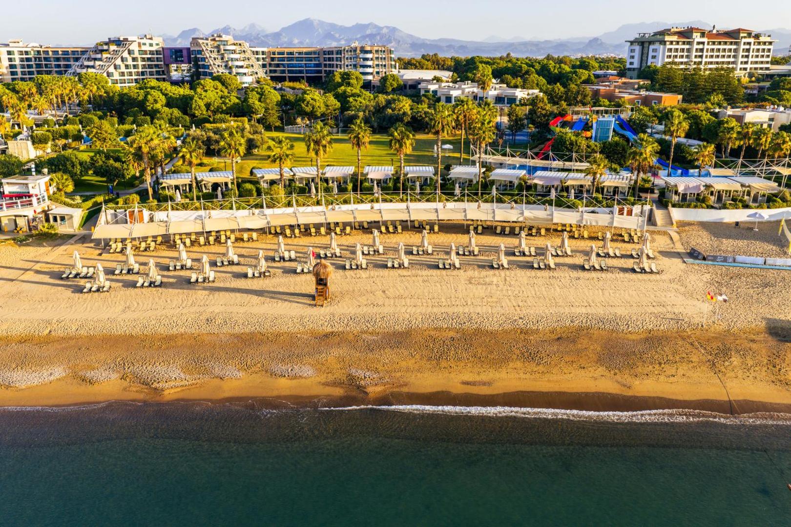 Susesi Luxury Resort privātā smilšu pludmale Belekā ar saulessargiem, sauļošanās krēsliem un skatu uz kalniem tālumā saulrieta gaismā