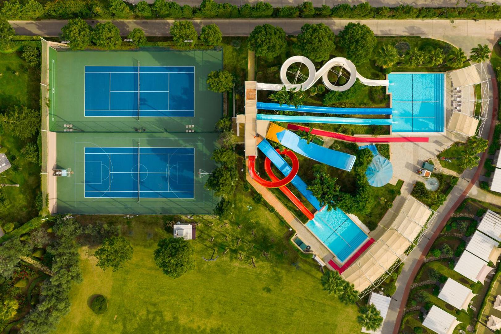 Susesi Luxury Resort sporta un izklaides zona no augšas — tenisa korti un krāsaini ūdens slidkalniņi Belekā