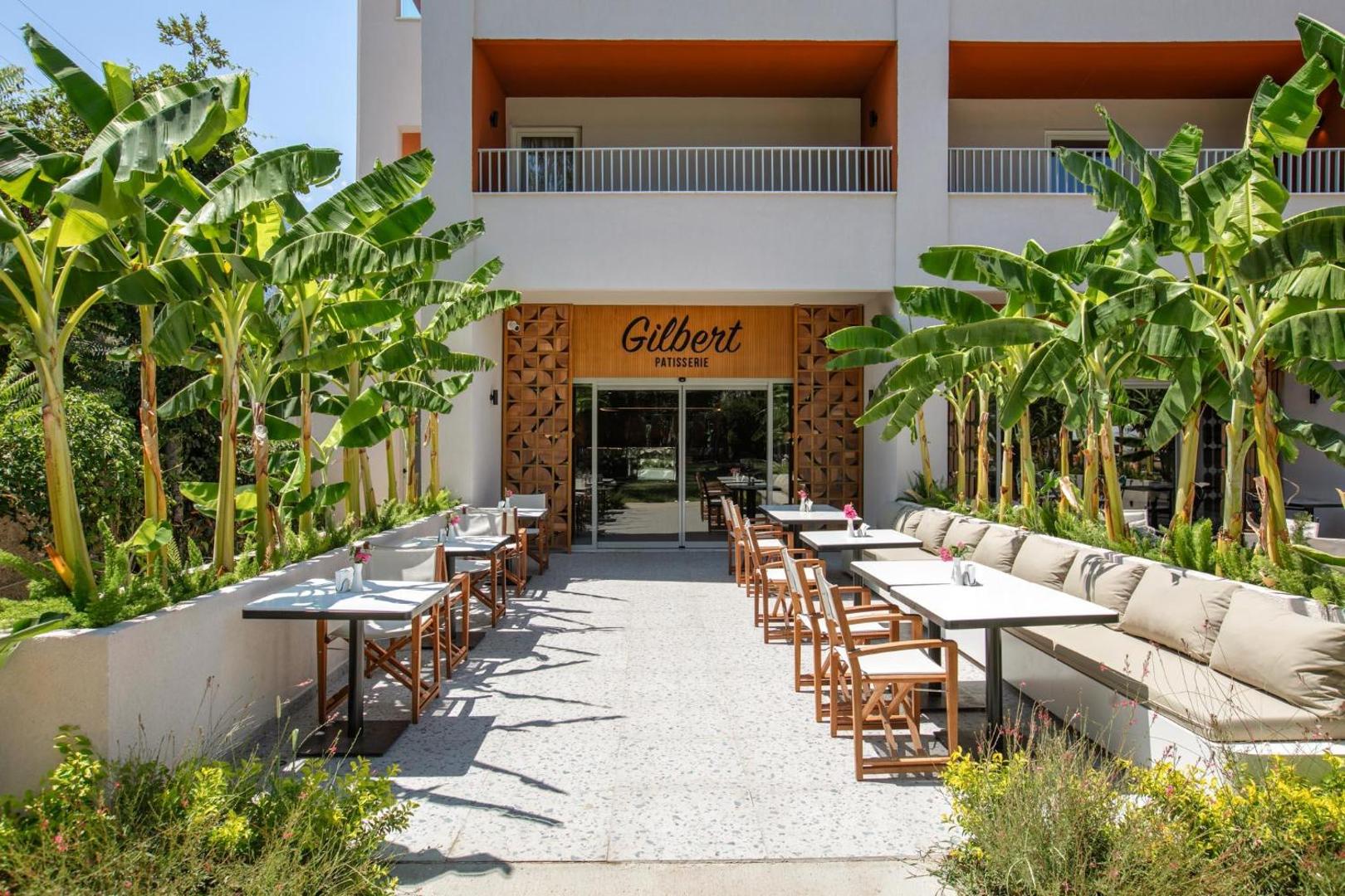 Greenwood Kemer Resort konditorejas Gilbert Patisserie āra zona ar baltiem galdiņiem un saulainu terasi banānu koku pavēnī Kemeras kūrortā