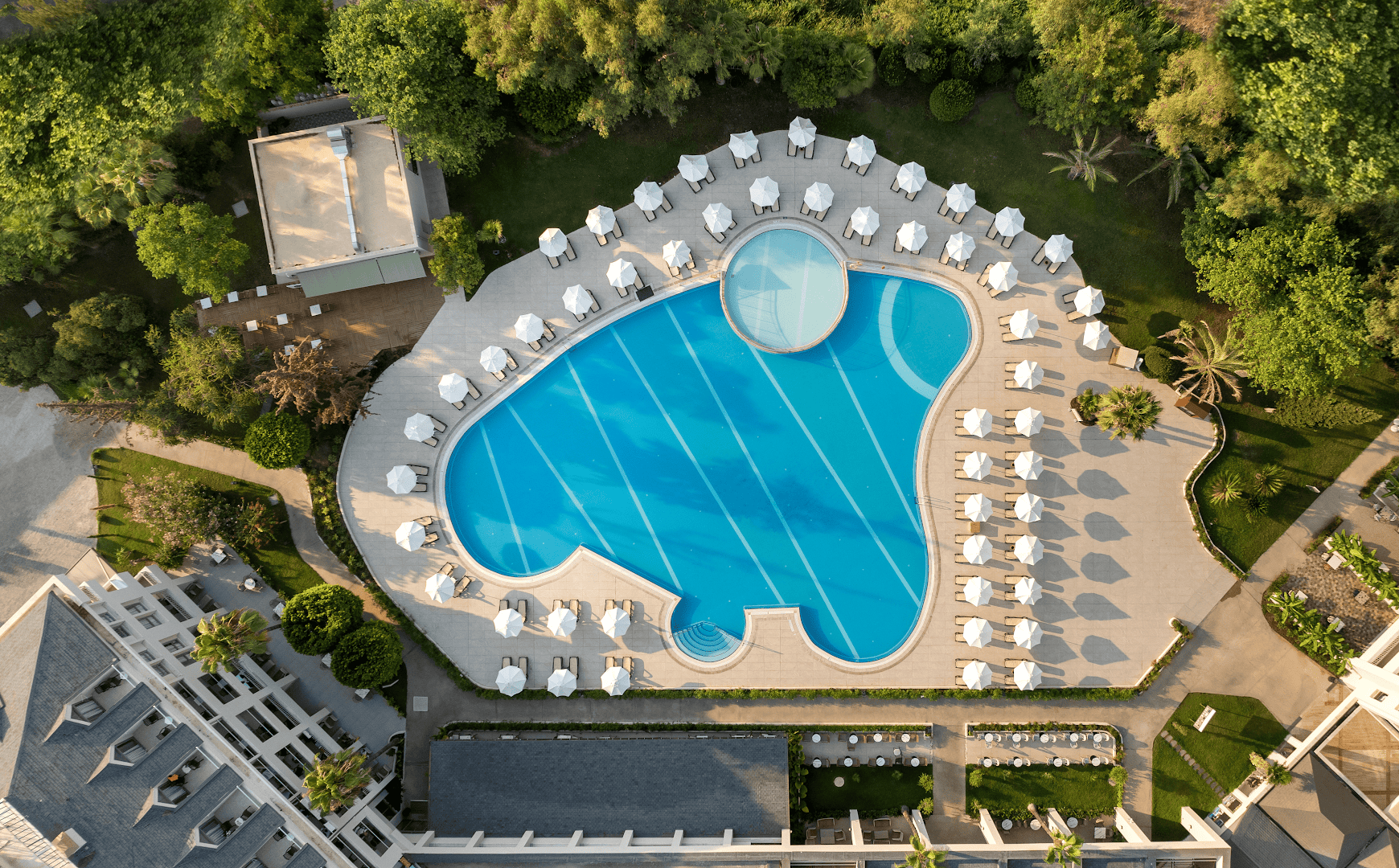 Greenwood Kemer Resort galvenā baseina skats no augšas ar saulessargiem, sauļošanās krēsliem un apkārtējo zaļo dārzu Turcijā.