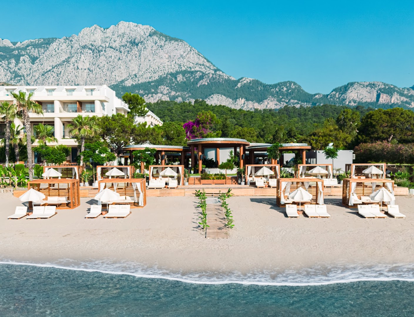 Sherwood Exclusive Kemer viesnīcas ēka un smilšu pludmale ar koka bungalo uz majestātisku kalnu fona Kemerā