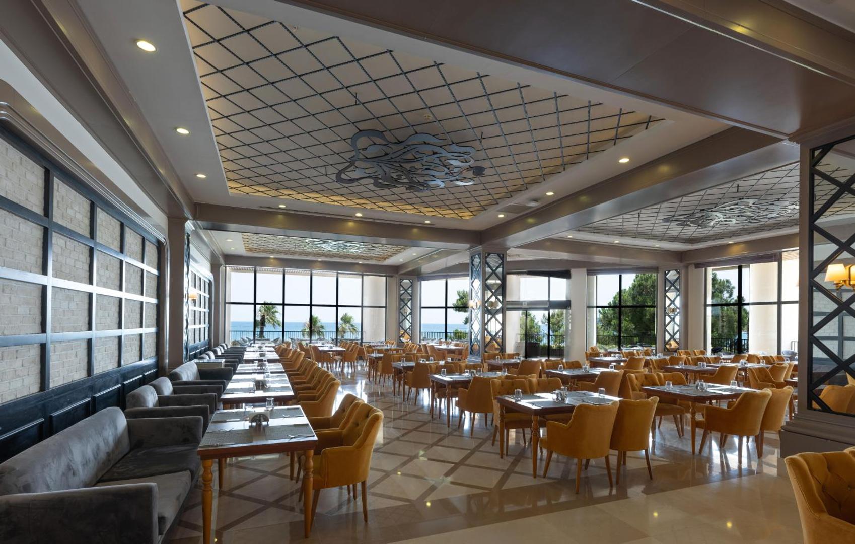 Sherwood Exclusive Kemer iekštelpu restorāns ar lieliem logiem, dzelteniem krēsliem un skatu uz dārzu un Vidusjūru.