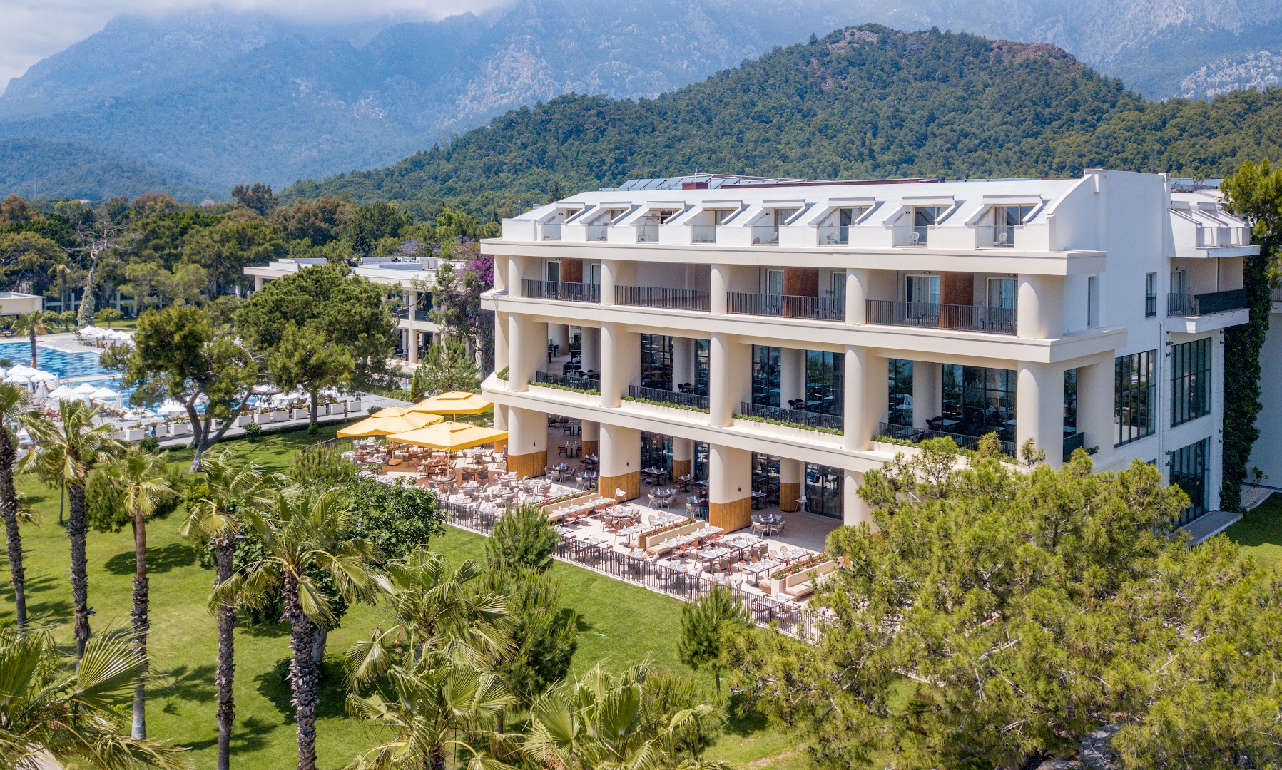 Viesnīcas Sherwood Exclusive Kemer arhitektūra ar restorāna zonu, palmām un skatu uz kalnu grēdu tālumā