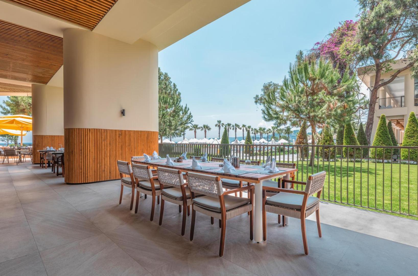 Sherwood Exclusive Kemer restorāna āra terase ar koka galdiem un krēsliem, skatu uz dārzu un palmām