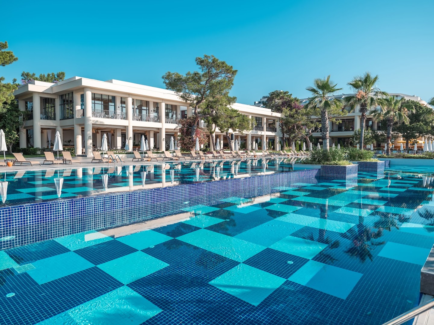 Sherwood Exclusive Kemer galvenais baseins ar ziliem flīžu rakstiem un viesnīcas ēku fonā — saulaina terase ar atpūtas krēsliem