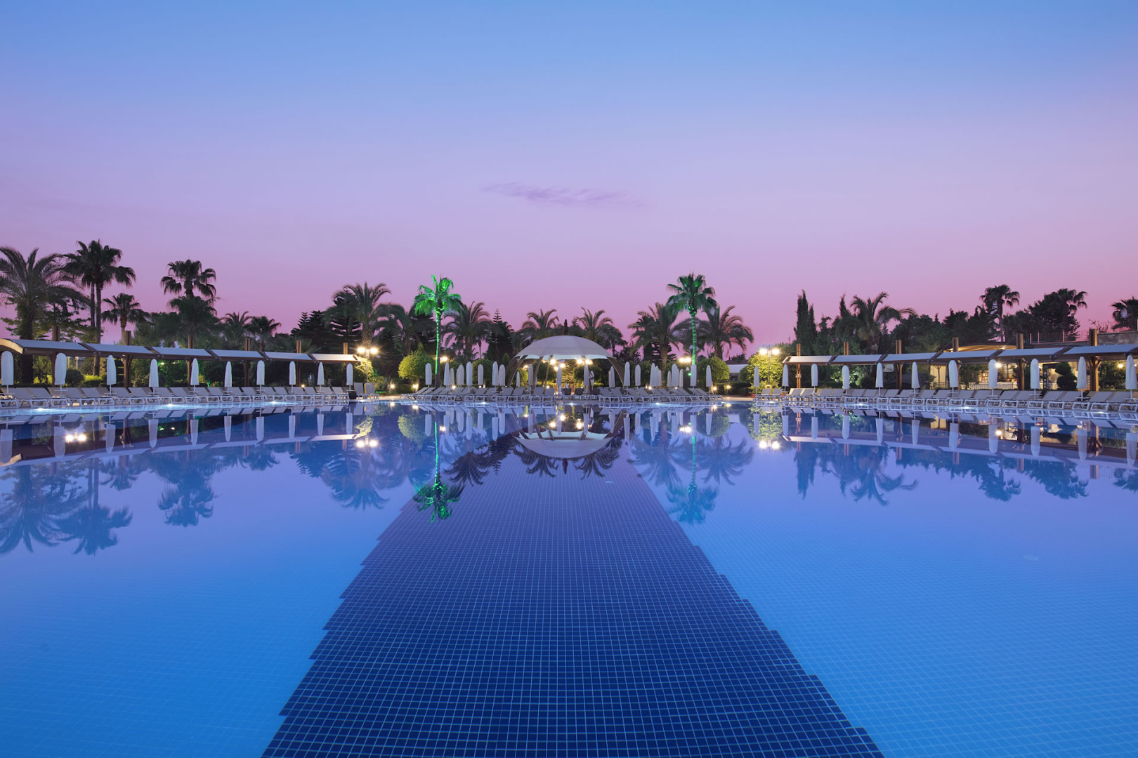 Saphir Resort & Spa lielais āra baseins krēslas stundā Alanijā — izgaismotas palmas un saulrieta debesis atspīd mierīgā ūdens virsmā