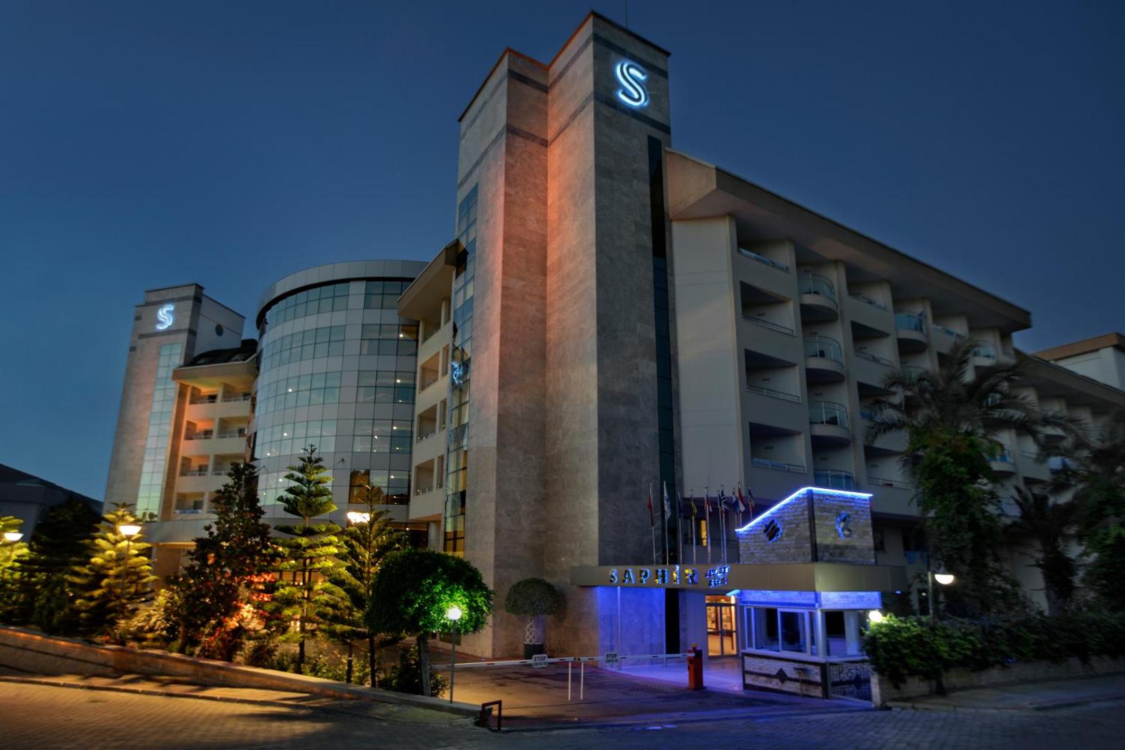 Saphir Resort & Spa galvenā ieeja un fasāde krēslas stundā — moderna daudzstāvu viesnīcas ēka ar logo un apstādījumiem