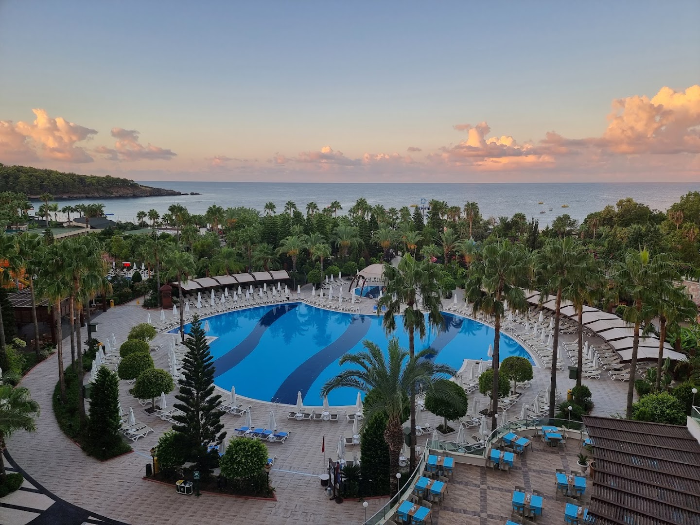 Panorāmas skats uz Saphir Resort & Spa baseinu, āra terasi un jūras piekrasti saulrieta laikā — mākoņi krāsojas siltos toņos