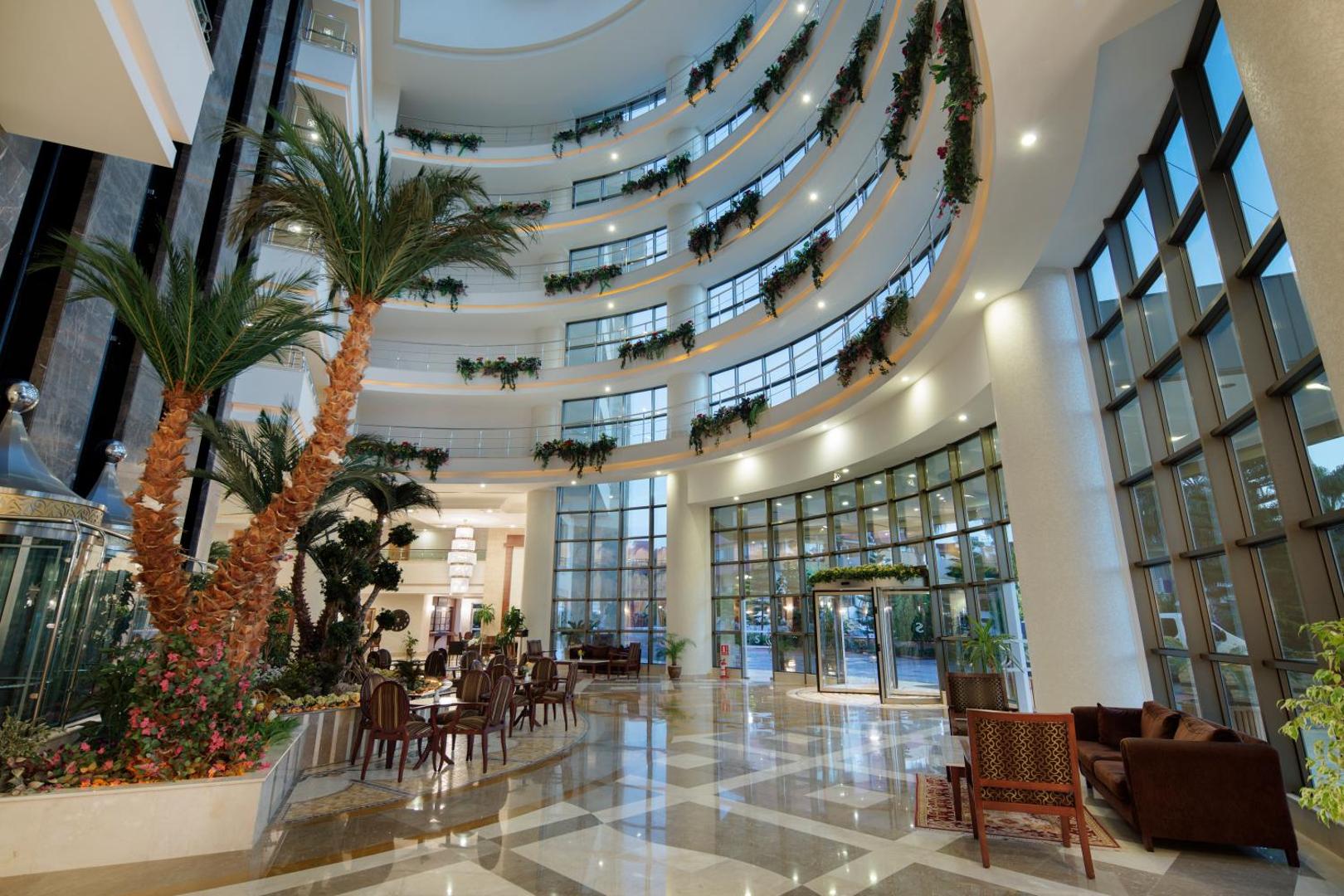 Saphir Resort & Spa plašais vestibils ar augstiem griestiem, palmām un atpūtas zonu Turcijas piekrastē — gaišs interjers ar panorāmas logiem