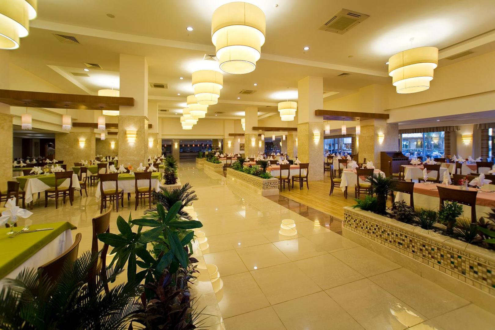 Saphir Resort & Spa plašā restorāna zāle ar siltu apgaismojumu, dekoratīviem augiem un klātiem galdiem Alānijas reģionā