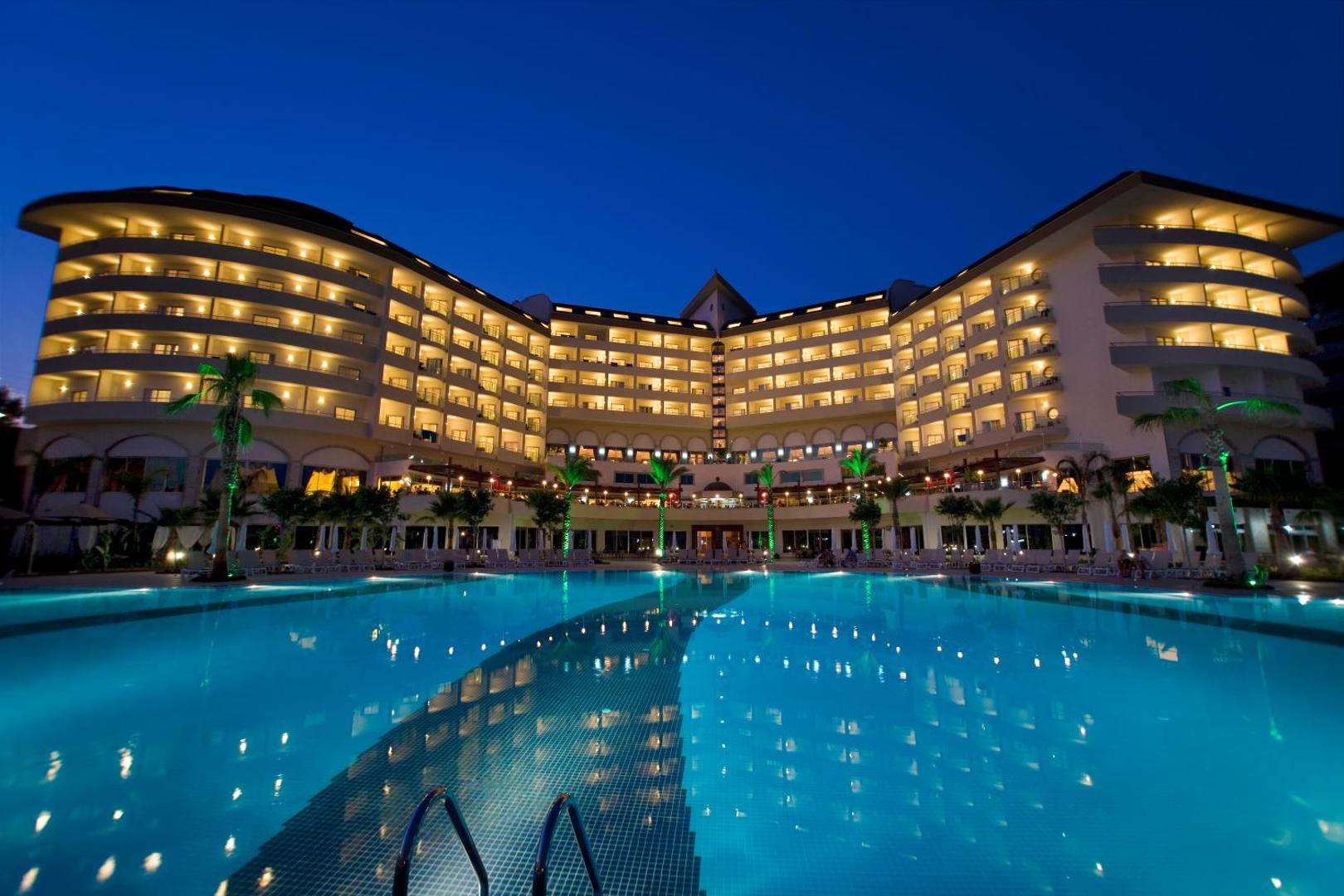 Saphir Resort & Spa viesnīcas ēka un izgaismots āra baseins vakarā Alānijā, Turcijā — moderna arhitektūra ar balkoniem un palmām