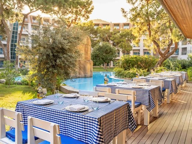 Rixos Sungate restorāna āra terase ar klātiem galdiem pie baseina — zili balti rūtaini galdauti un skats uz viesnīcas dārzu