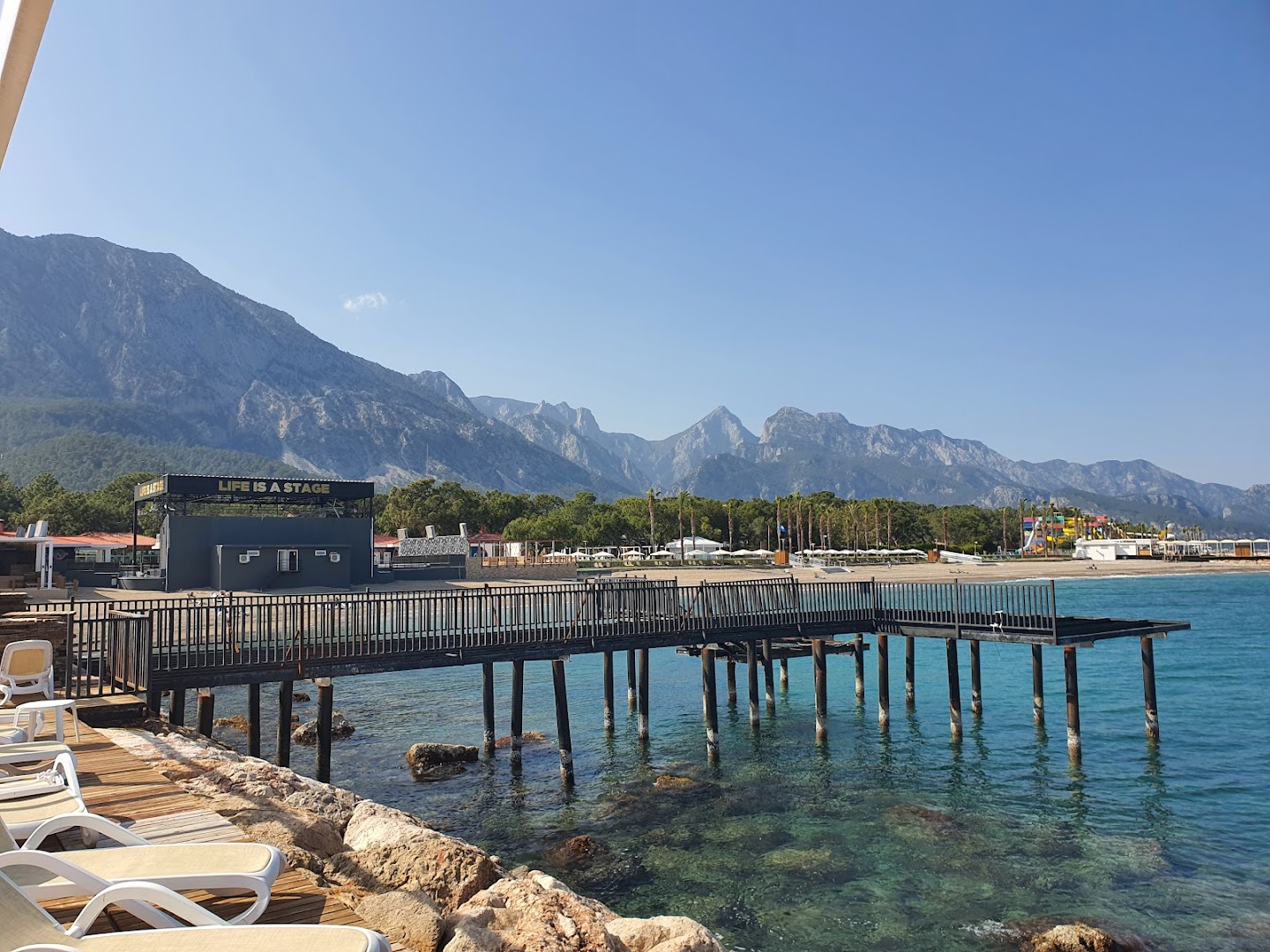 Rixos Sungate skats no piestātnes uz kalniem un pludmales skatuvi Kemeras reģionā — caurspīdīgs ūdens un akmeņaina piekraste