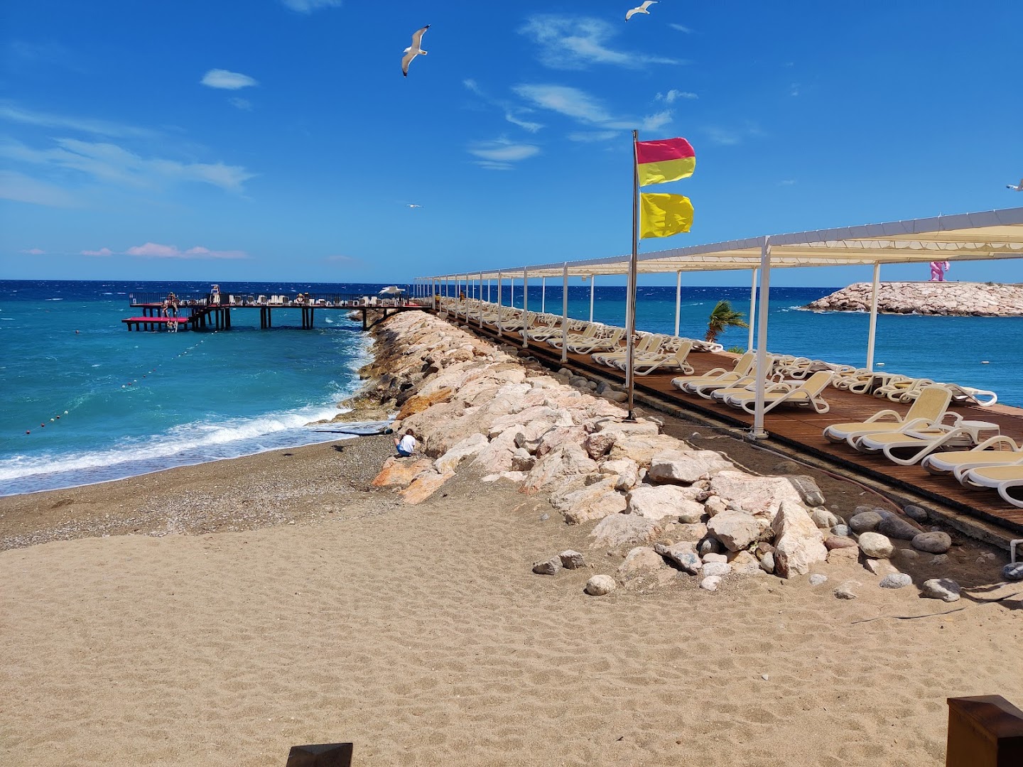 Rixos Sungate smilšu pludmale ar sauļošanās krēsliem zem nojumēm un piestātni tālumā — skats uz zilo Vidusjūru saulainā dienā