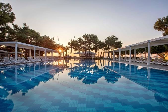 Saulriets virs Rixos Sungate lielā āra baseina ar simetriskiem sauļošanās krēsliem un priežu meža siluetiem.