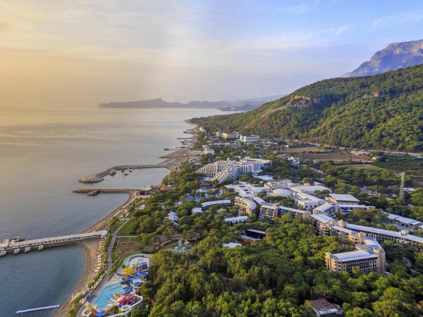 Rixos Sungate panorāmas skats no putna lidojuma uz viesnīcas kompleksu, piekrasti un kalniem Beldibi reģionā, Turcijā saulrieta laikā