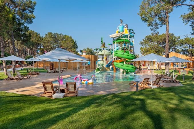 Rixos Sungate bērnu baseins ar krāsainiem slidkalniņiem un ūdens atrakcijām — sauļošanās krēsli un lietussargi zālienā zem priedēm