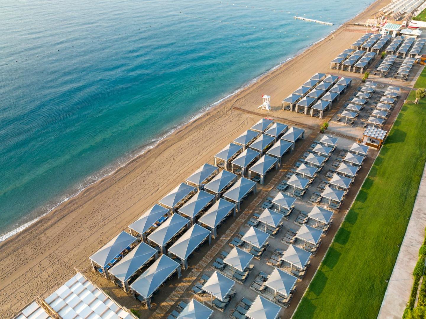 Rixos Premium Tekirova pludmales rinda ar modernām nojumēm un sauļošanās krēsliem pie dzidra jūras ūdens