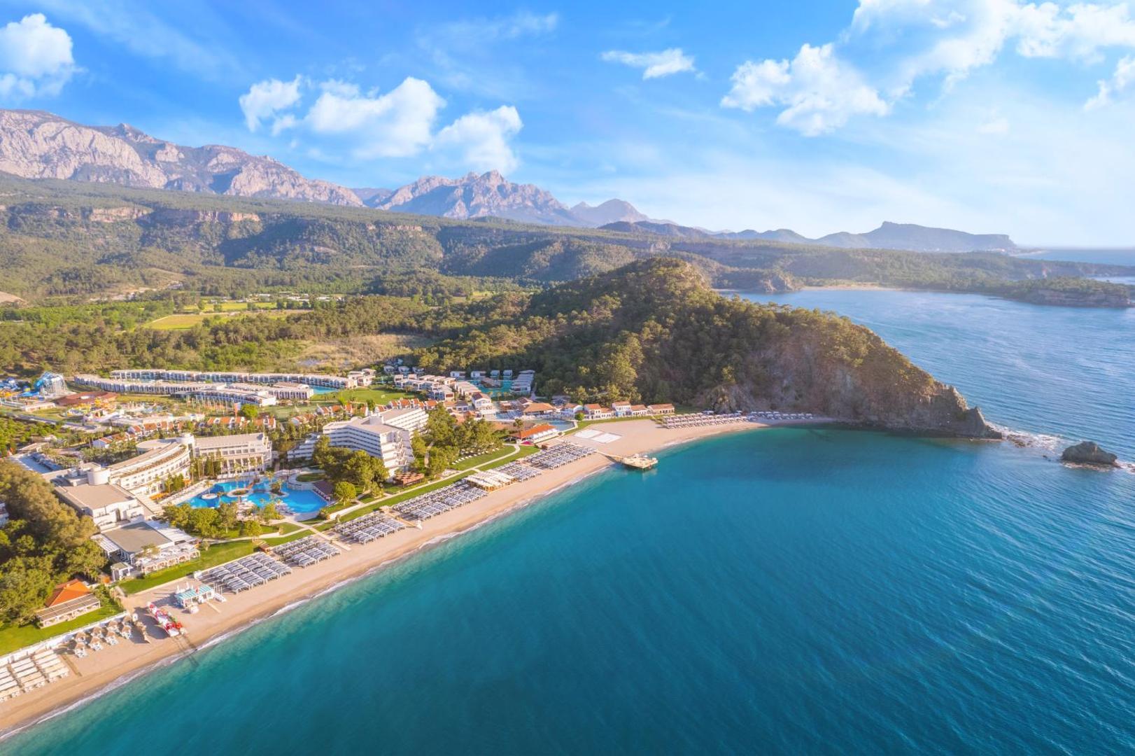 Rixos Premium Tekirova ainava ar viesnīcas ēkām, smilšu pludmali un zilajiem kalniem saulainā dienā