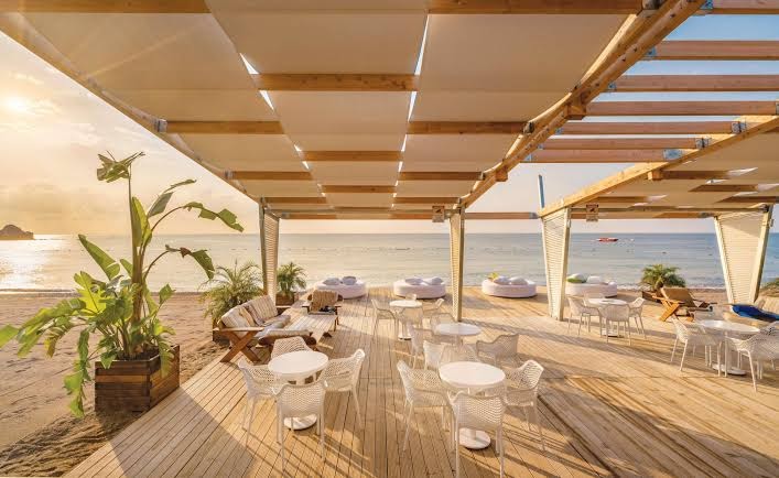 Rixos Premium Tekirova pludmales bārs un atpūtas zona ar koka terasi, baltām mēbelēm un skatu uz Vidusjūru