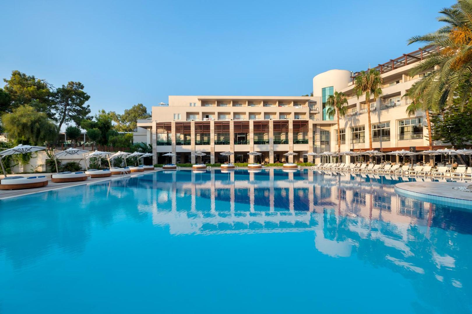 Rixos Premium Tekirova galvenā ēka un lielais āra baseins dienas laikā — gaiša fasāde, saulessargi un palmas piekrastē